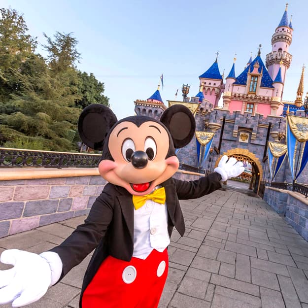Mickey Mouse in a tuxedo waving in front of Sleeping Beauty Castle.
