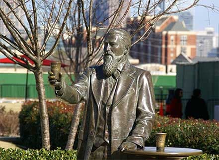 Bronze statue of a bearded man holding a glass, with city buildings in background.