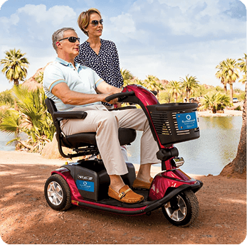 Couple enjoying a park, man driving a red mobility scooter, woman standing beside him.