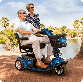 Man on a blue mobility scooter, woman standing behind him by a lake with palm trees.