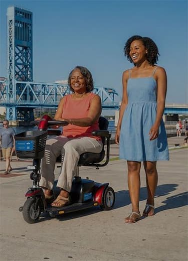 Two women, one in a mobility scooter, smile on a sunny city riverwalk.