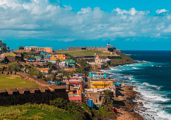 Colorful coastal town with a historic fort overlooking the blue ocean under a partly cloudy sky.